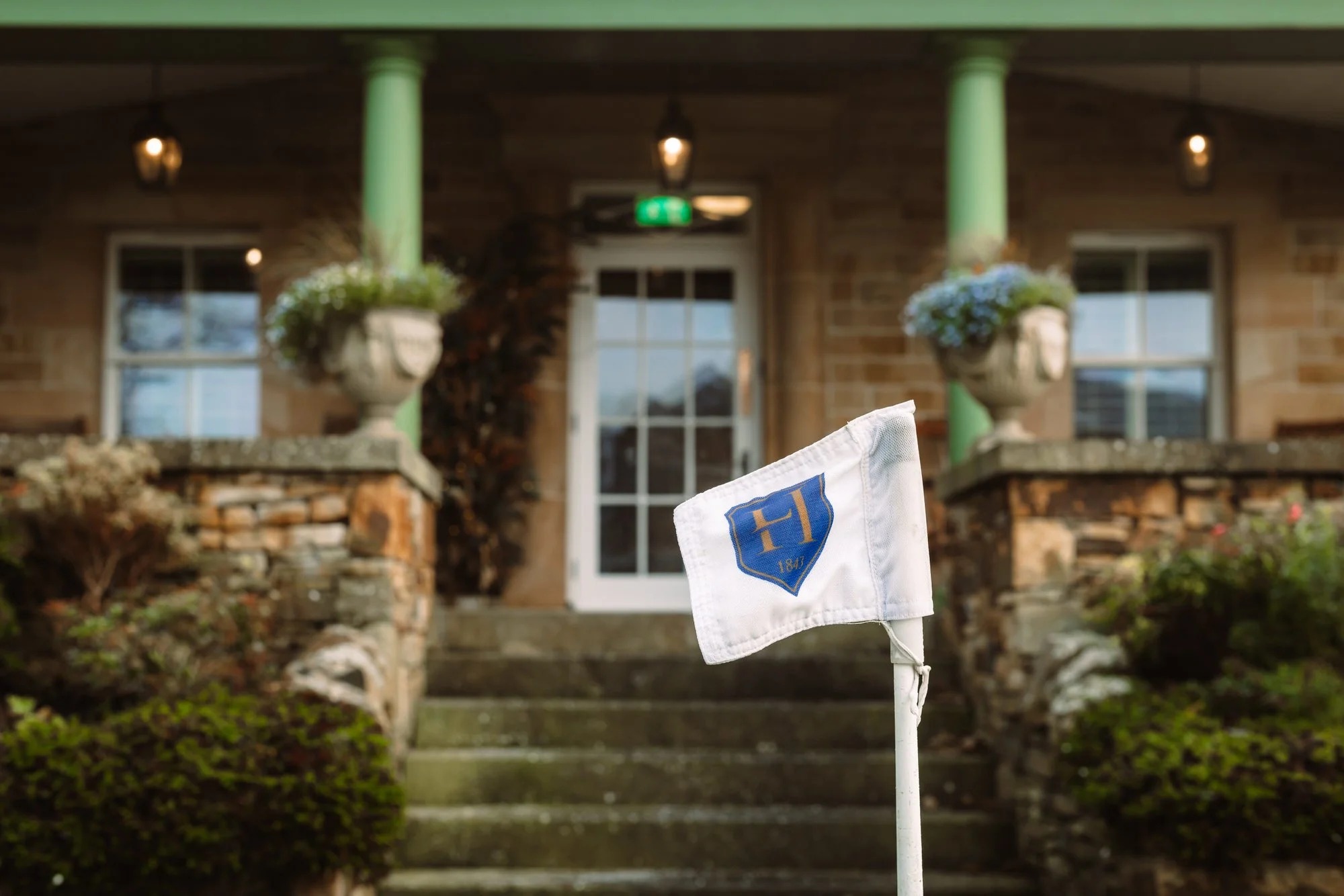 A monogrammed golf flag stands before a colonnaded stone porch with hydrangeas in urns either side of the entrance.