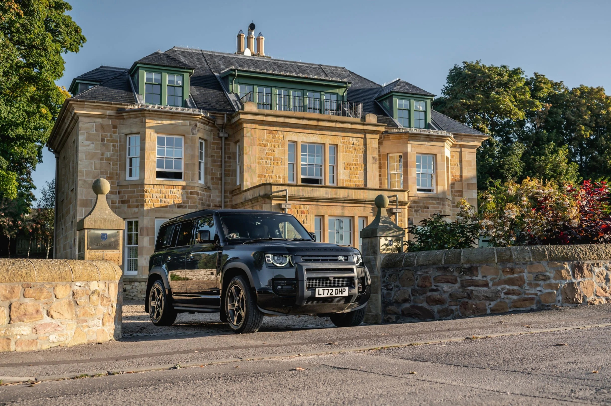 A black 4x4 is parked before a grand stone mansion with a green conservatory roof, gateposts topped with stone finials either side.