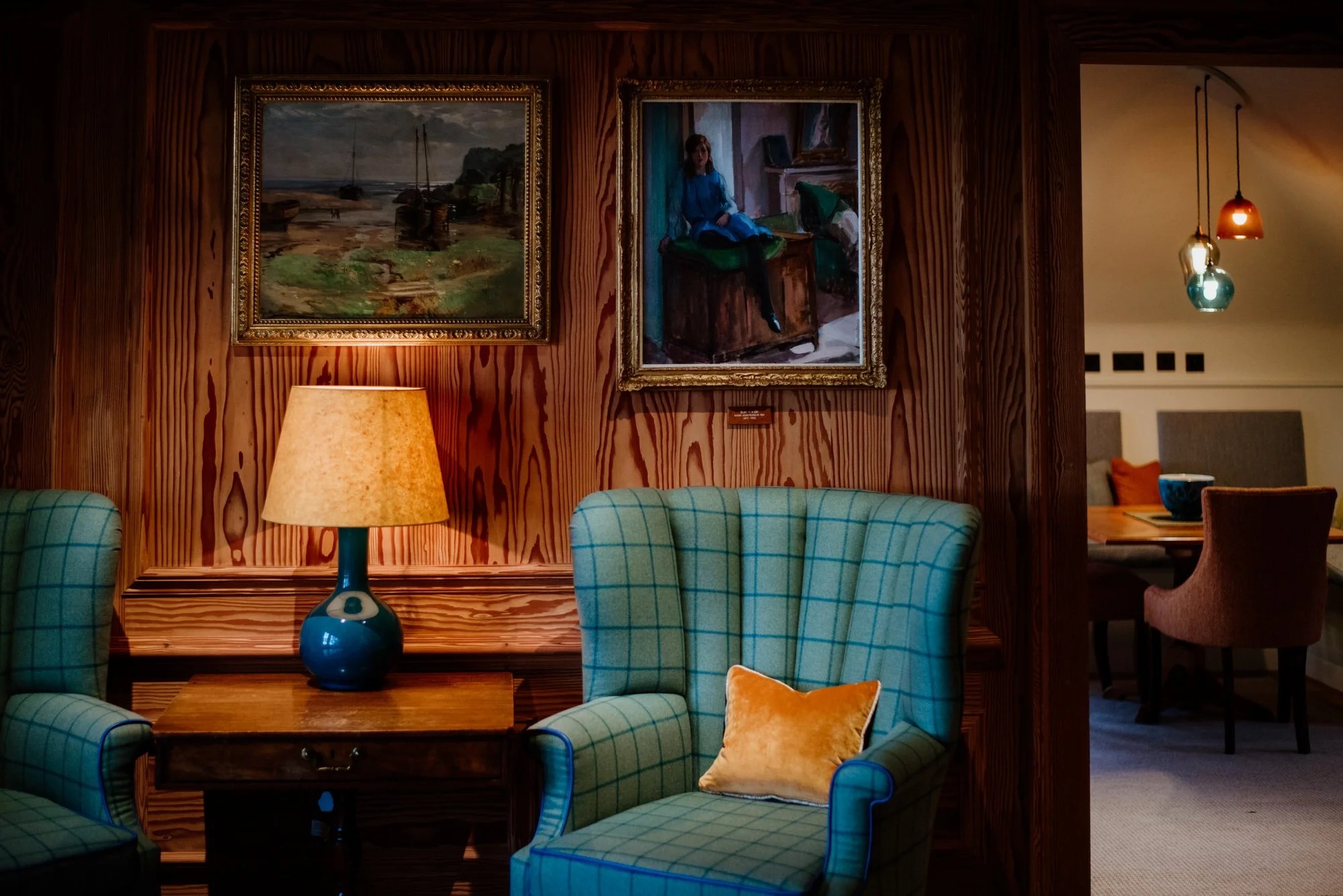 Two teal check armchairs flank a lit lamp table beneath framed paintings, a doorway opening onto a dining room beyond.