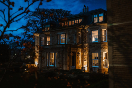 A grand stone mansion glows with lit windows at dusk, bare autumn branches framing the facade.