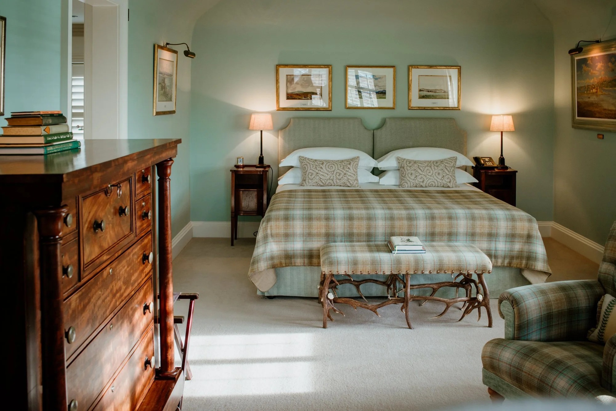 A mahogany chest of drawers stands beside a plaid-covered bed with a footstool at its end, three framed prints above the headboard.