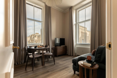 A round table and chairs stand before tall curtained windows looking onto stone buildings, a sofa and mounted television to the right.