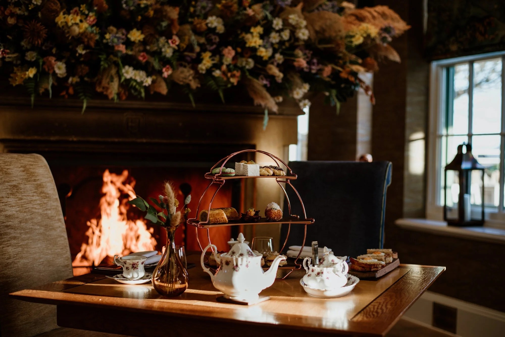 A silver cake stand of pastries stands beside a teapot and cups on a table before a lit fireplace decked with an autumnal floral display.