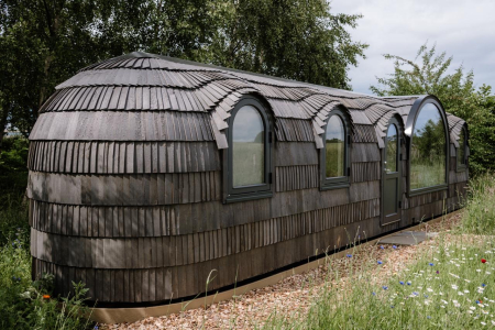 A curved cabin clad in overlapping wooden shingles stands on gravel among wildflowers, arched windows and a door along one side.