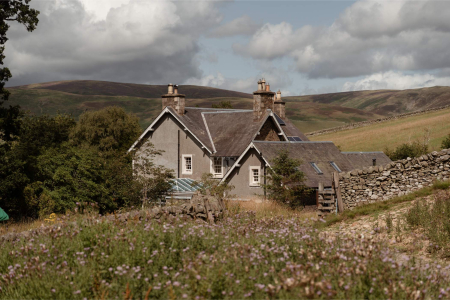 A grey stone farmhouse with several chimneys stands against rolling hills, a wildflower meadow in the foreground.