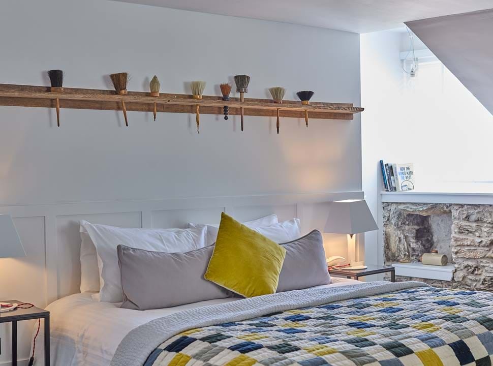 A row of paintbrushes is displayed on a shelf above a bed dressed with grey pillows and a mustard cushion, a stone fireplace to the right.