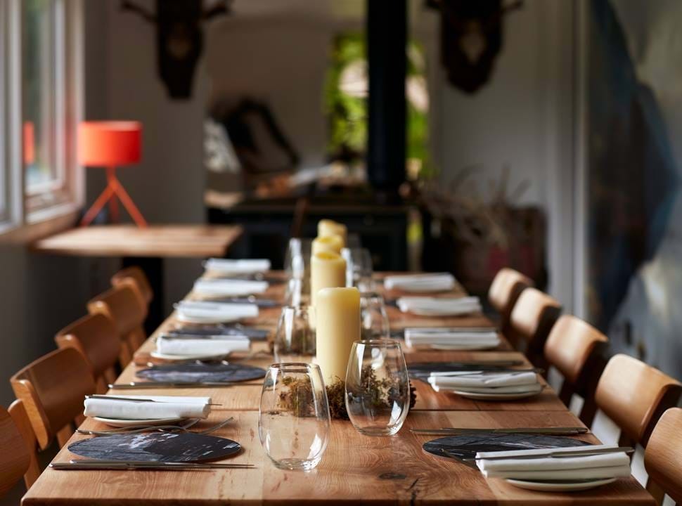 A long table set for a formal dinner with candles and black placemats stretches between wooden chairs, antlers mounted on the wall behind.