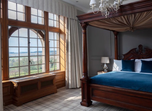 A four-poster bed with a dark wood frame and blue linen sits beside arched windows looking onto a green landscape.