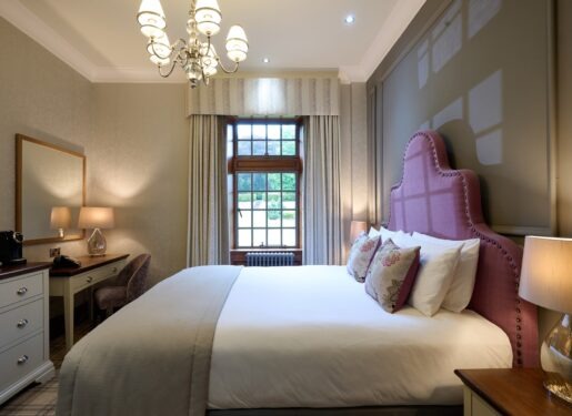 A pink upholstered headboard sits beneath a chandelier, a dressing table and mirror to the left and a curtained window behind.