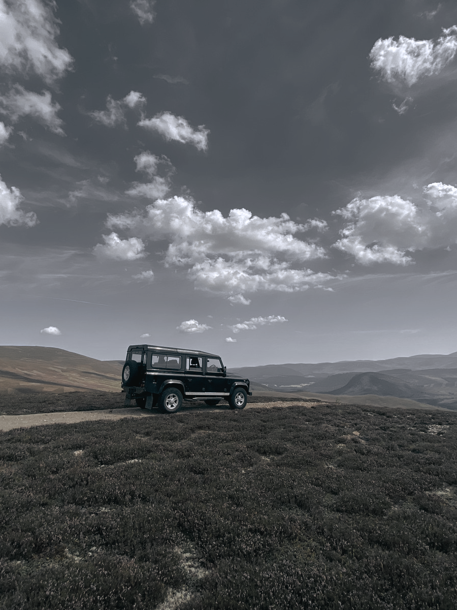 A black Land Rover Defender parked on a hill track beneath a sky scattered with clouds, distant hills stretching to the horizon.