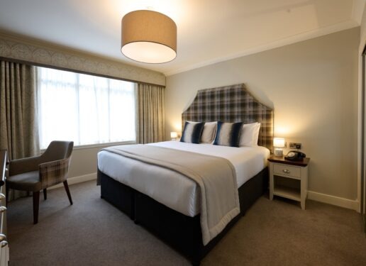 A bed with a tartan upholstered headboard sits beside a check armchair, curtains framing a bright window.