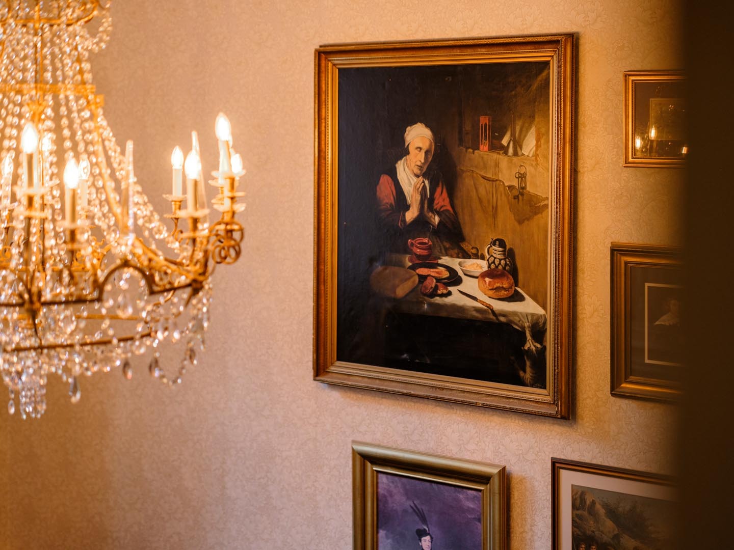 A gilt chandelier hangs beside a gallery of framed paintings on a patterned wall.