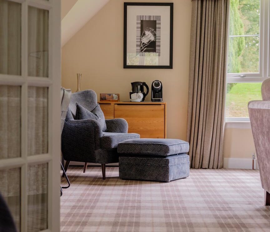 A grey armchair and matching footstool sit before a chest of drawers with a kettle and coffee machine, a framed print above and a curtained window to the right.