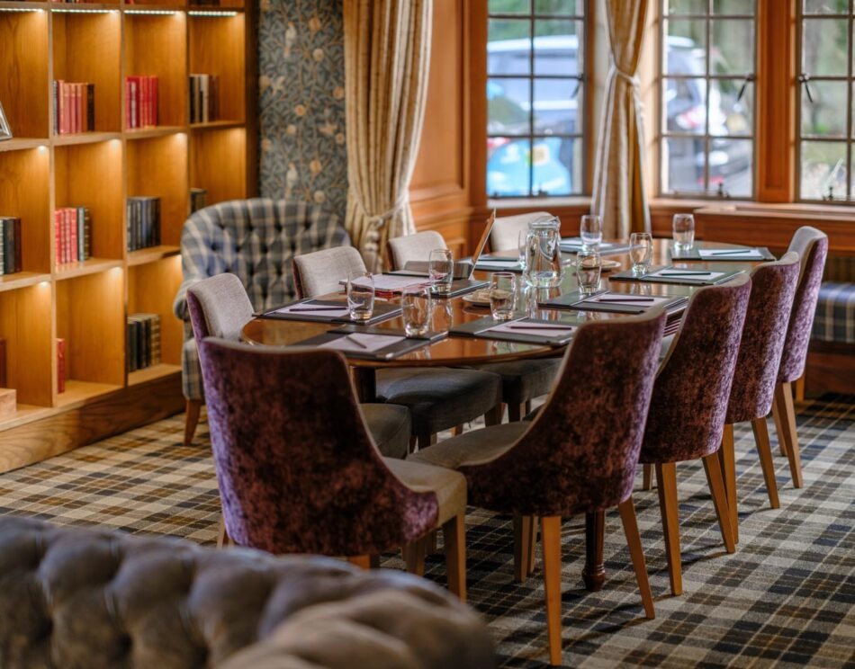 A long dining table set with glasses and menus stands before built-in bookshelves, patterned armchairs and velvet dining chairs arranged around it.