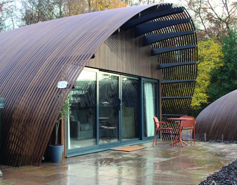 A curved timber-shingled building with glazed doors opens onto a paved patio, orange chairs and a table set outside.