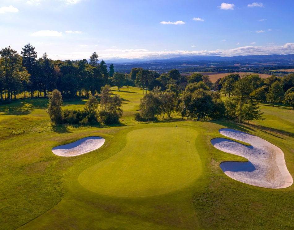 An aerial view of a golf green surrounded by bunkers and trees, rolling countryside stretching to the horizon.