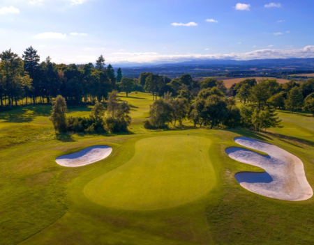 An aerial view of a golf green surrounded by bunkers and trees, rolling countryside stretching to the horizon.