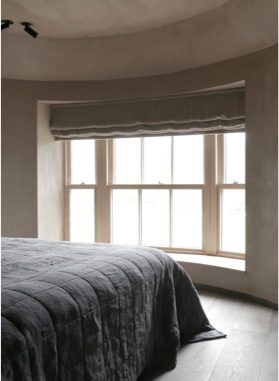 A grey quilted bedspread lies on a bed before a curved bay window with a pleated blind, morning light streaming in.