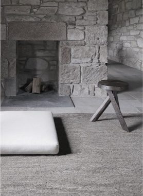 A pale cushion and a three-legged wooden stool sit on a woven rug before a rough stone fireplace with a small stack of logs.