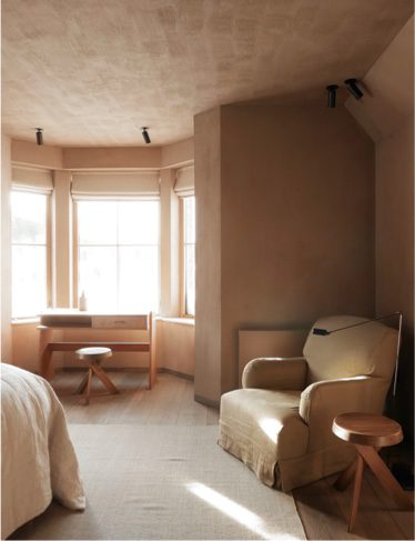A pale armchair and low stool sit in the corner of a warmly lit room, a writing desk and stool visible by the curtained bay window.