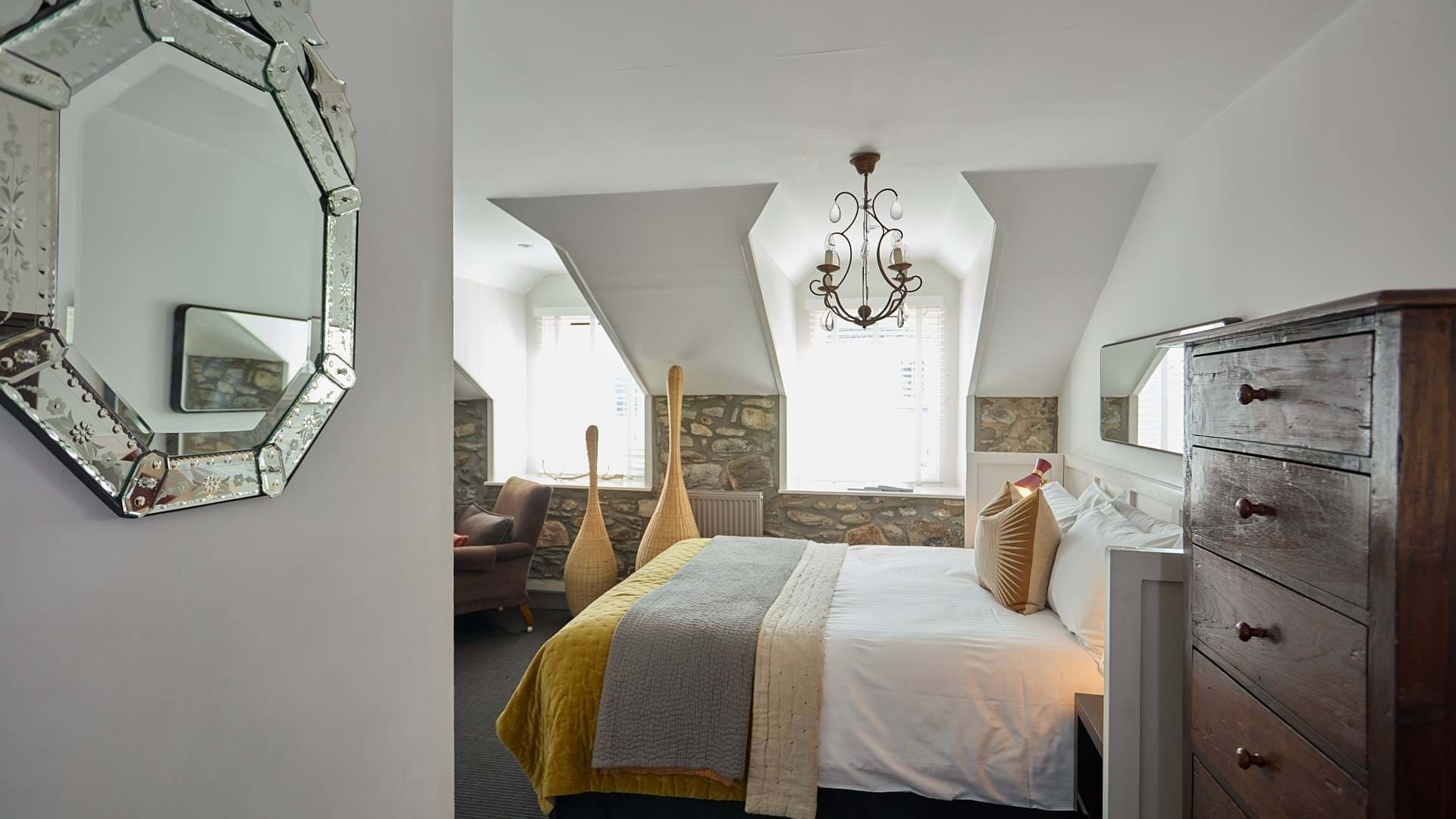 An octagonal mirror hangs beside a bed with a mustard throw, exposed stone walls and a chandelier under the sloped ceiling.