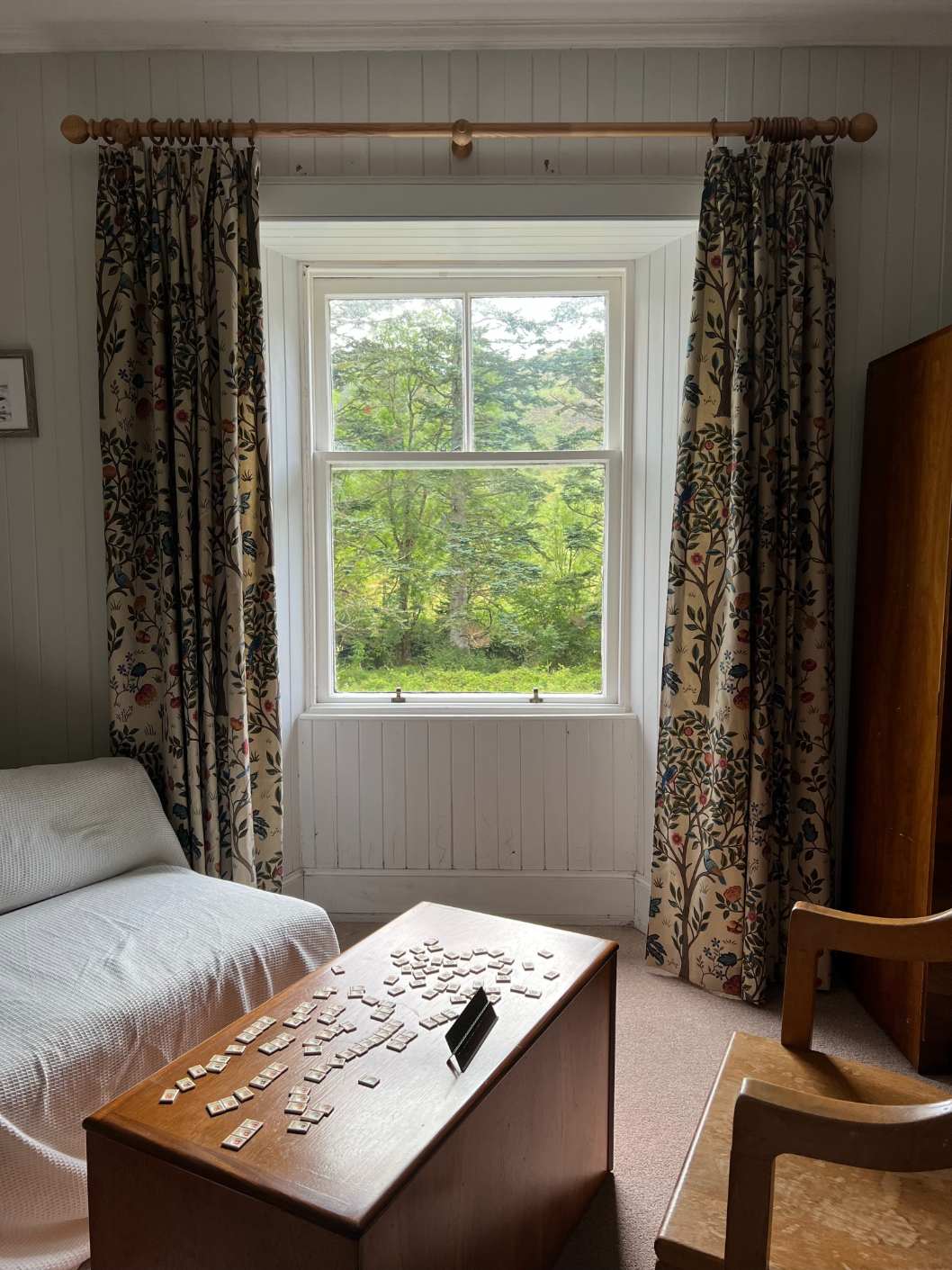A window dressed with floral curtains looks onto woodland, a wooden chest with letter tiles scattered across it in the foreground.