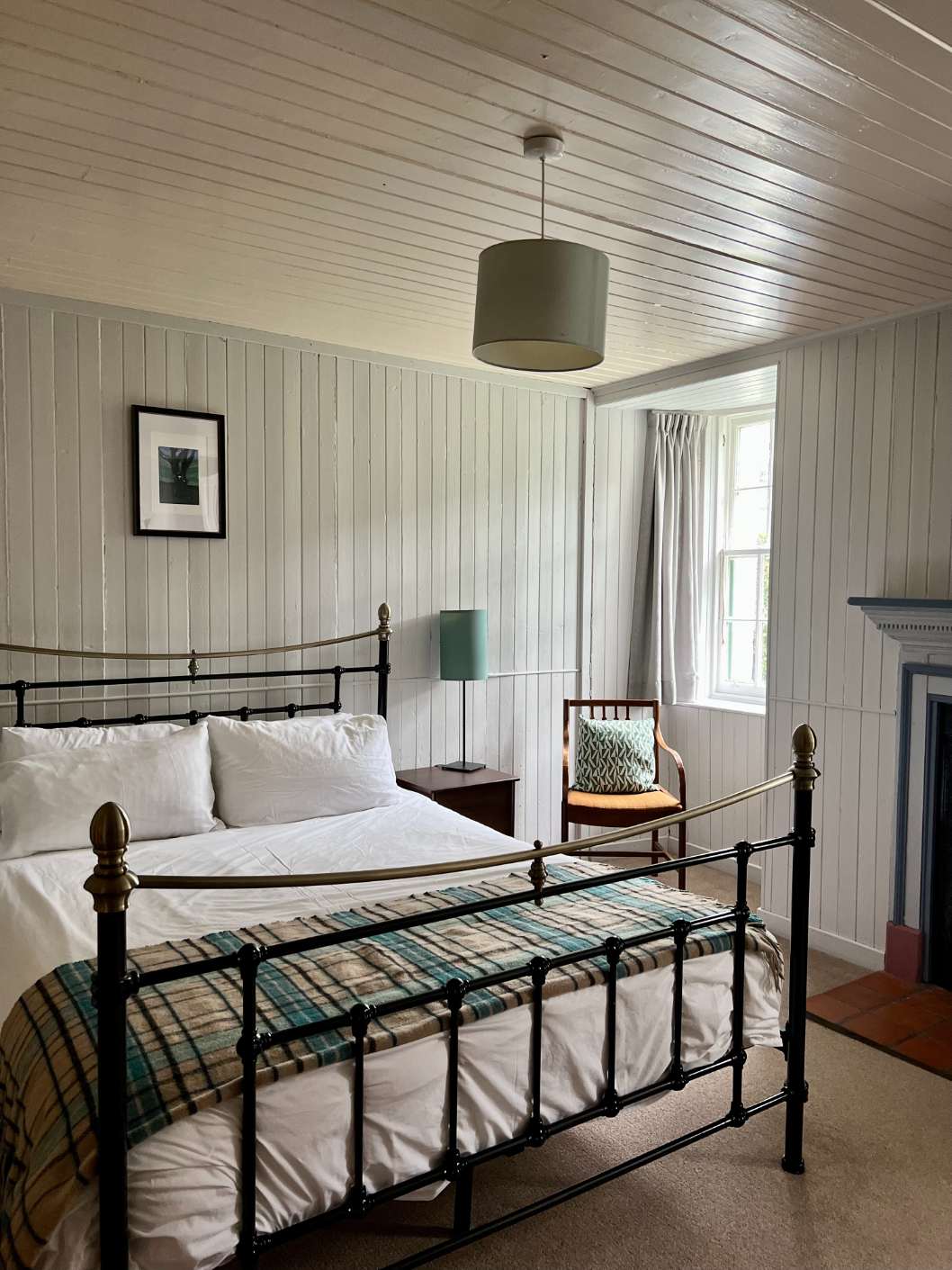 A brass bed with tartan bedding sits before a curtained window, a wing-back chair and old fireplace to the right of the room.