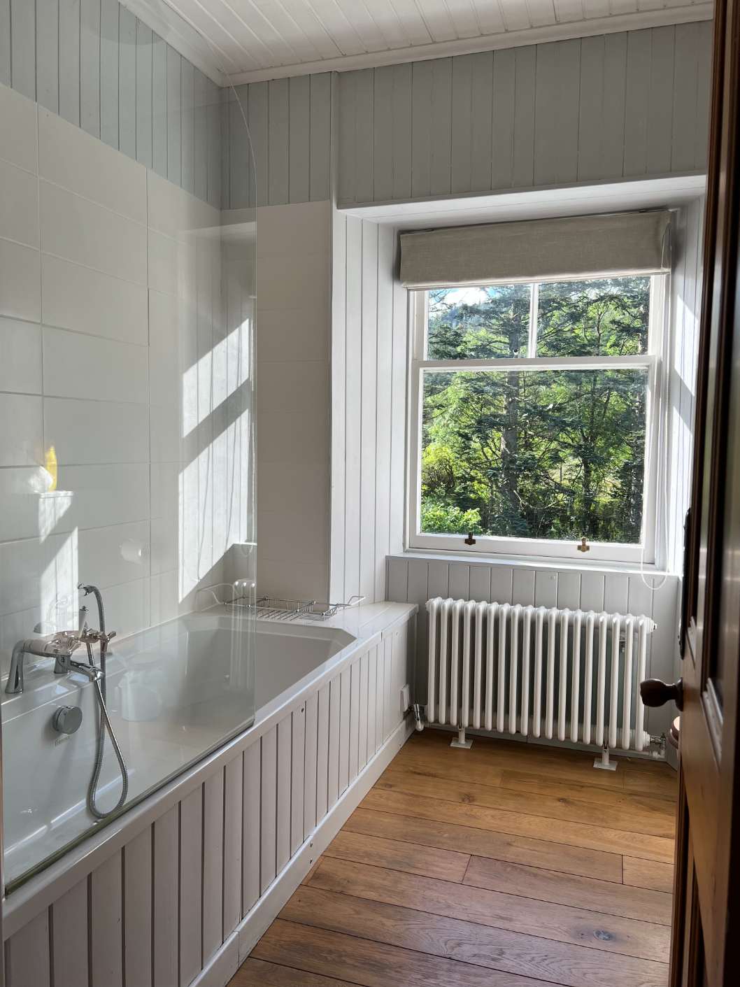A bath with a glass screen stands below a window looking onto trees, a radiator and wood panelling completing the room.