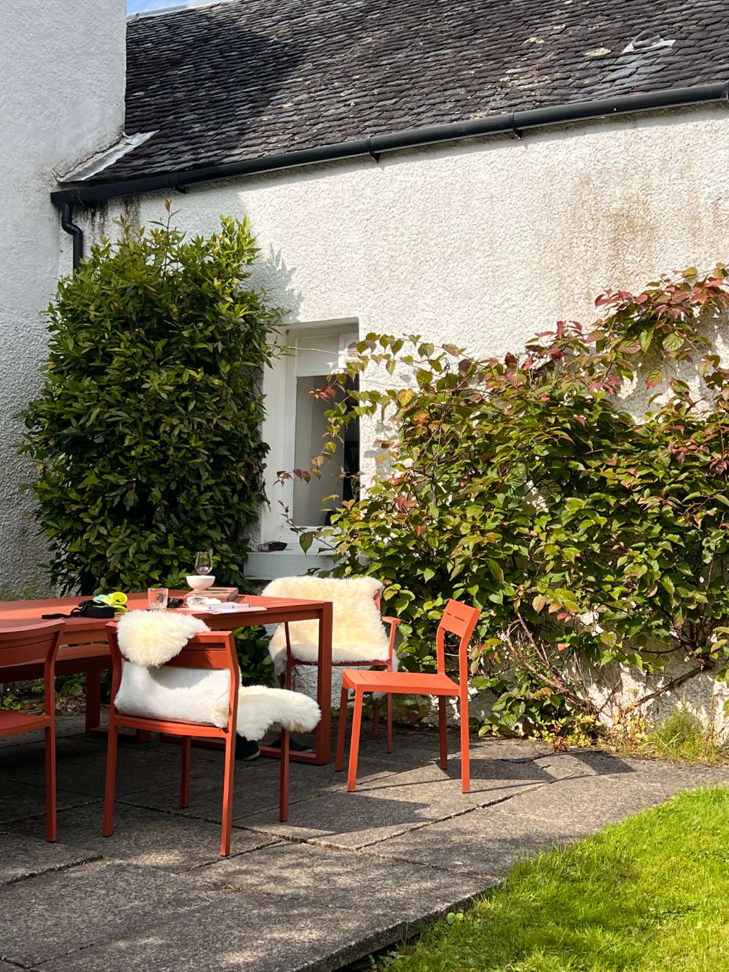 An orange table and chairs draped with sheepskins stand on a patio beside a white house, climbing shrubs along the wall.