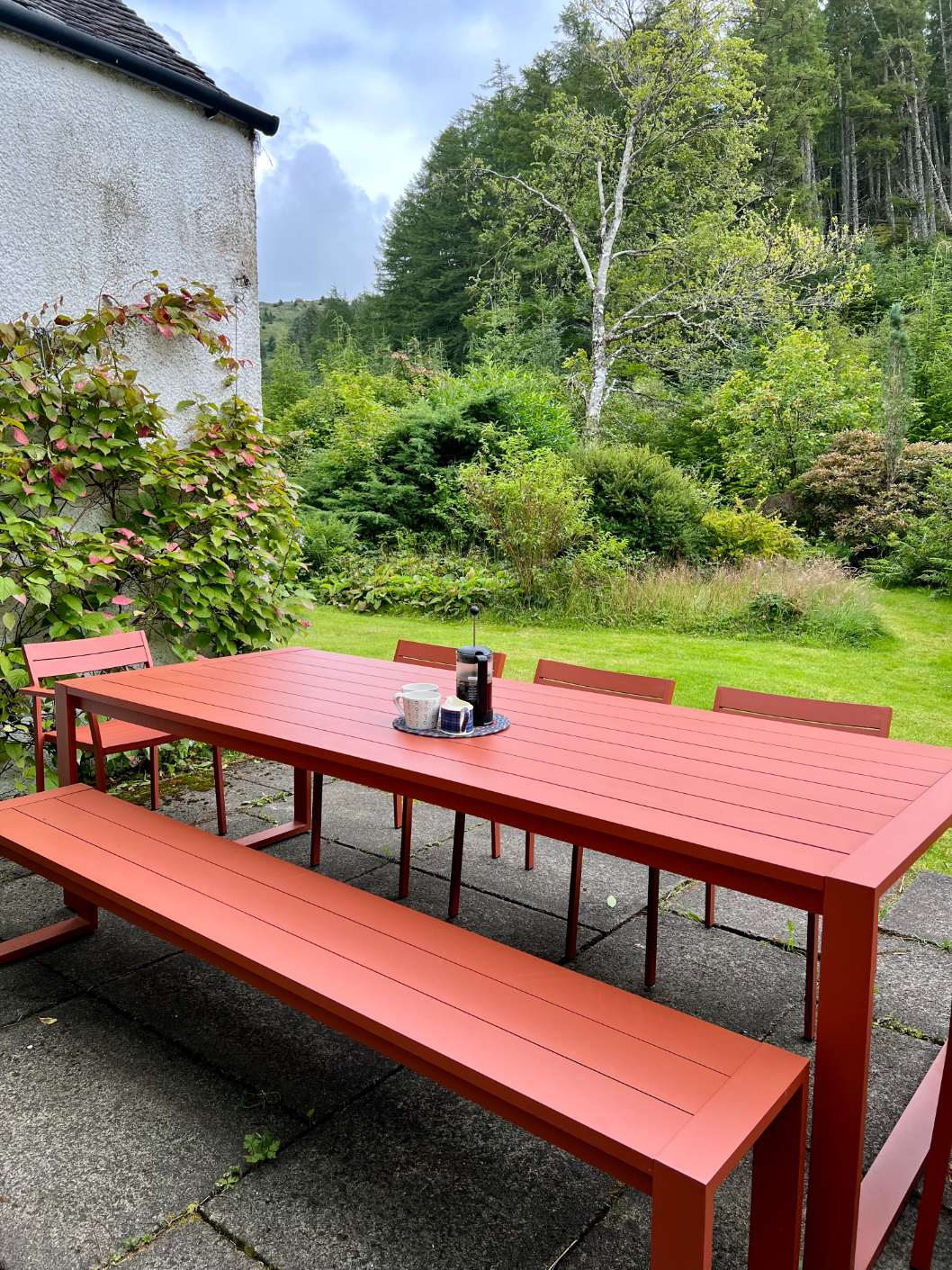 An orange dining table and bench stand on a patio before a white house, hills and trees visible beyond the lawn.