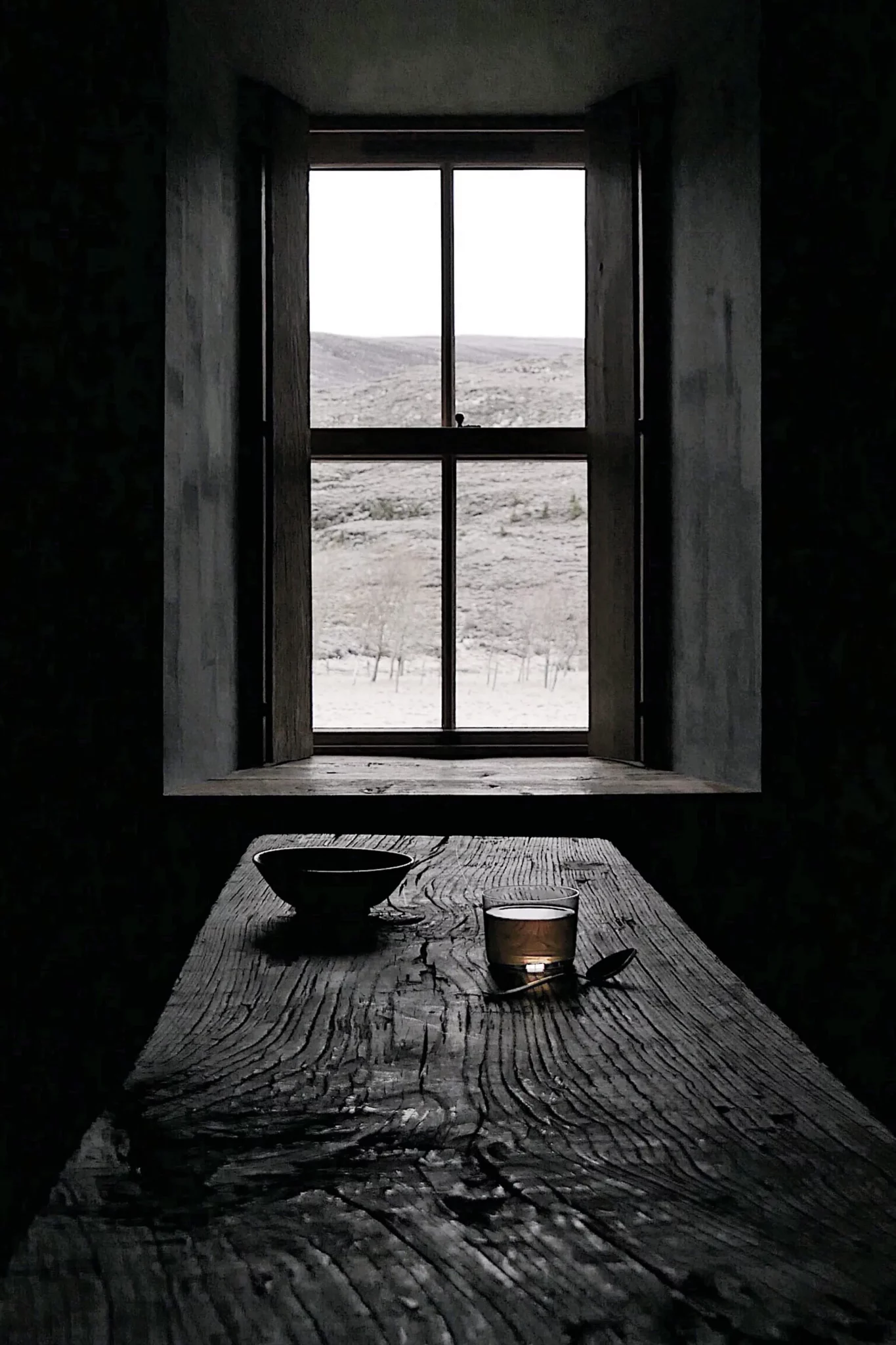 A bowl and a lit candle sit on a weathered wooden table before a window framing moorland.