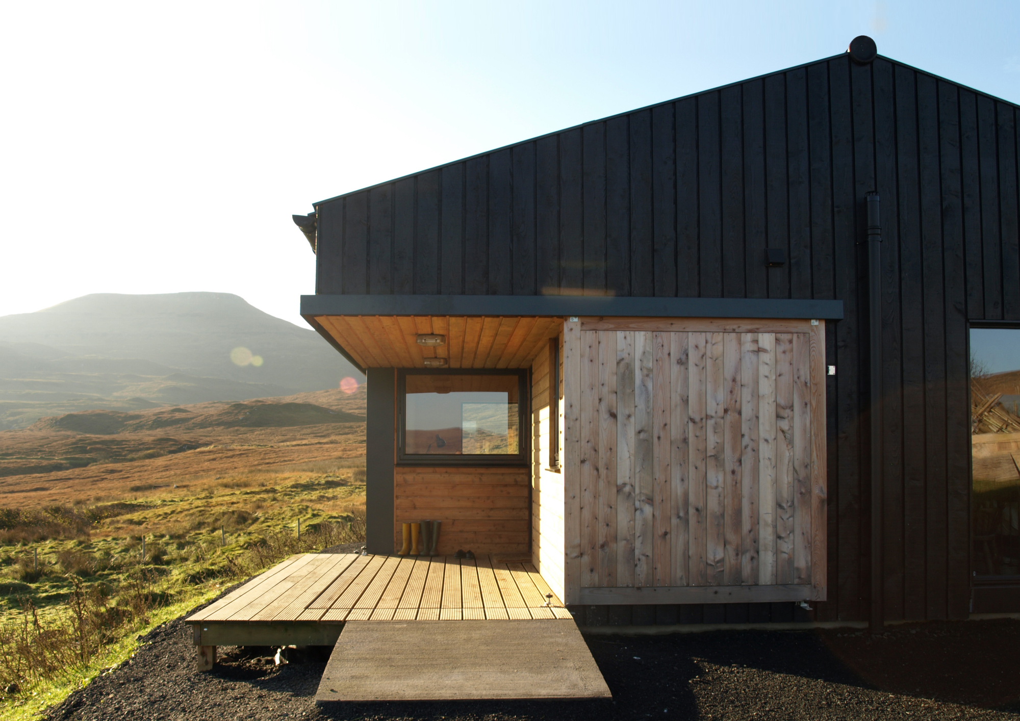 A black timber cabin with a sliding wooden door catches the low evening sun, moorland and hills stretching behind it.