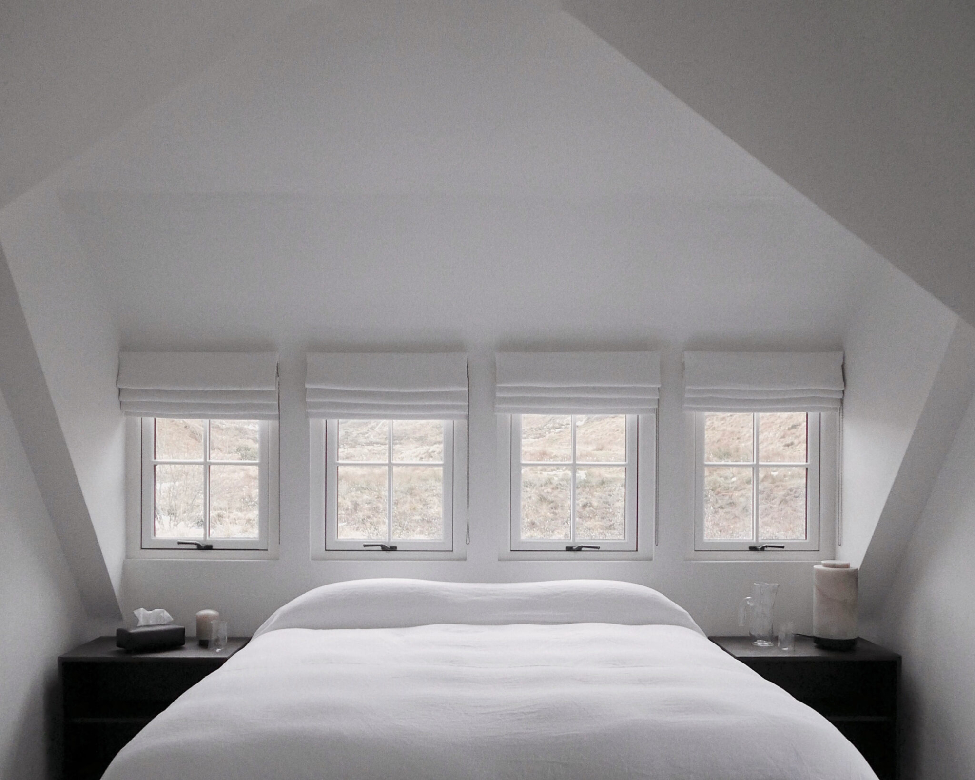A bed with white linen sits beneath four dormer windows fitted with roman blinds, bedside tables either side.