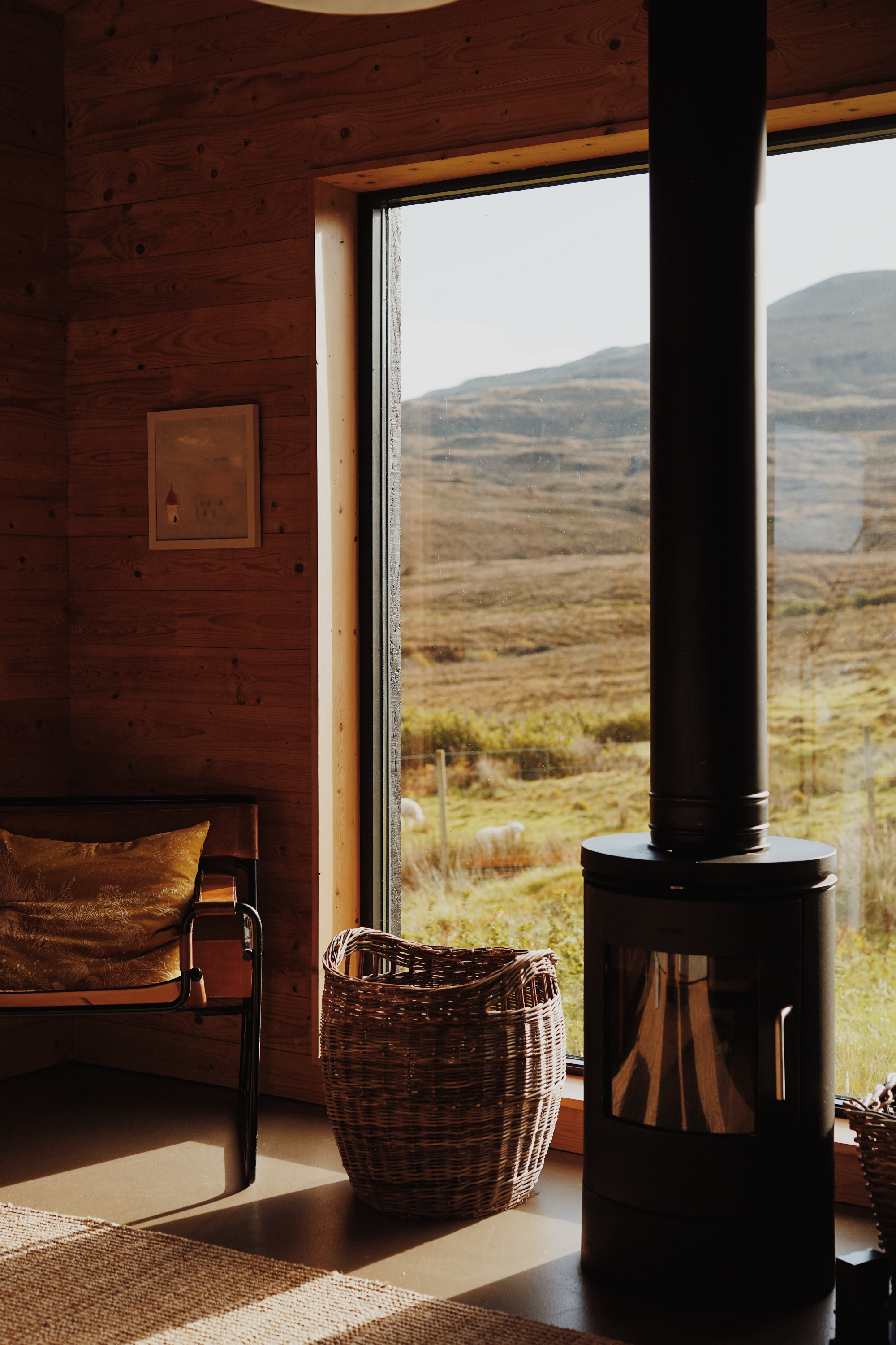 A leather sling chair with a mustard cushion sits beside a wicker basket, a wood burning stove set into the corner before a window onto moorland.