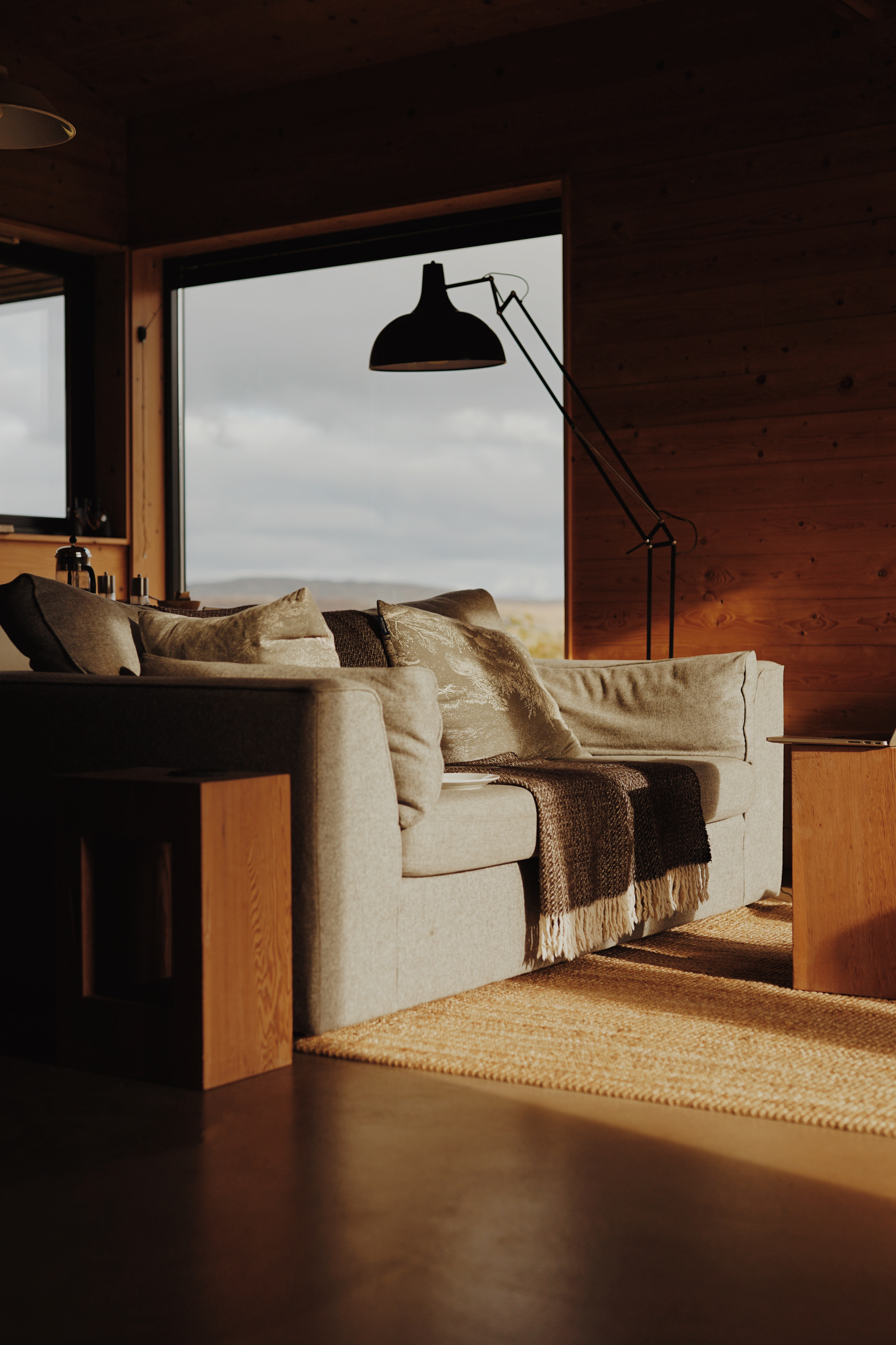 A beige corner sofa with cushions and a fringed throw sits before a window framing moorland, a black angled floor lamp beside it.