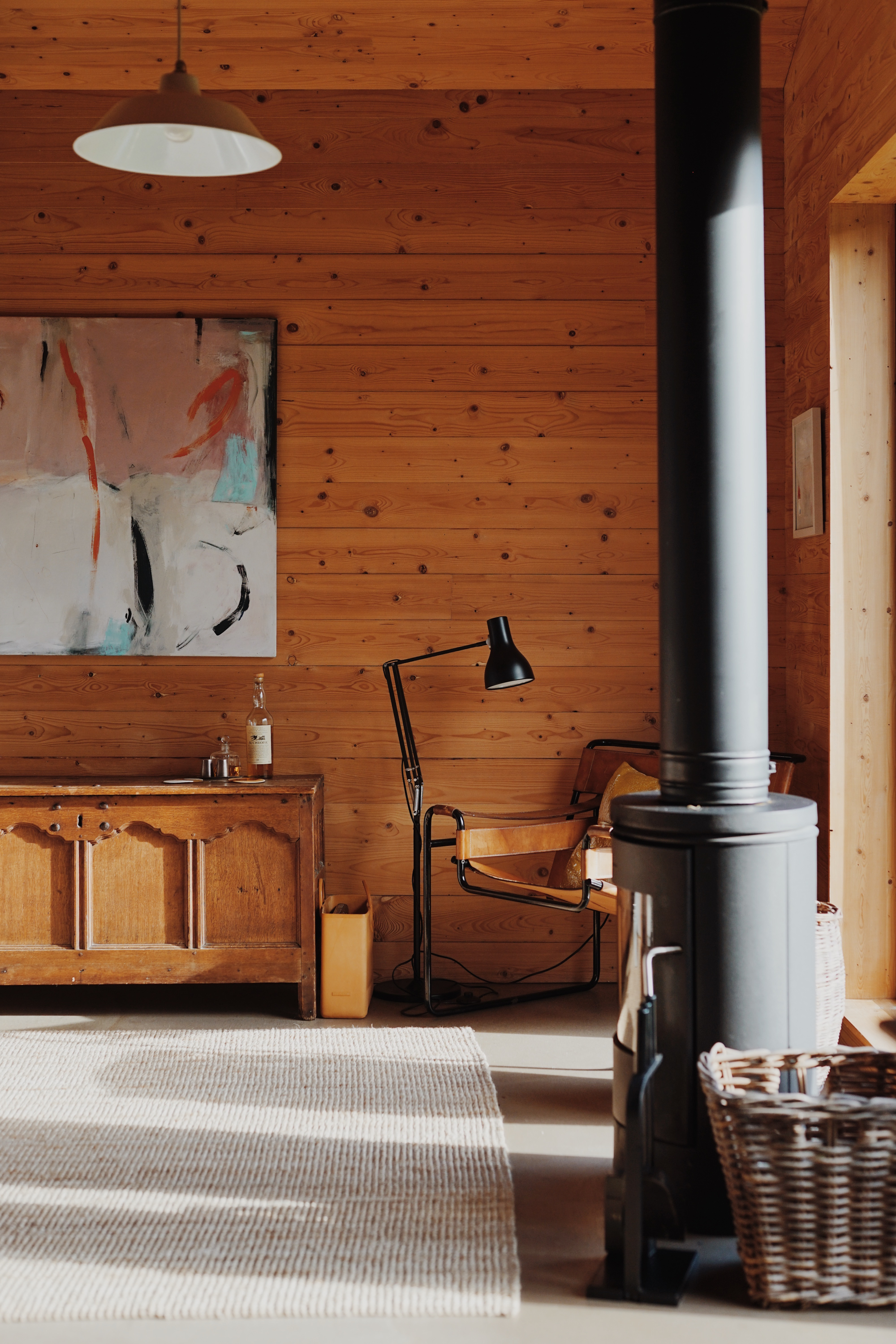 A leather sling chair with a mustard cushion sits beside a wicker log basket and wood burning stove, an abstract painting above an antique chest.