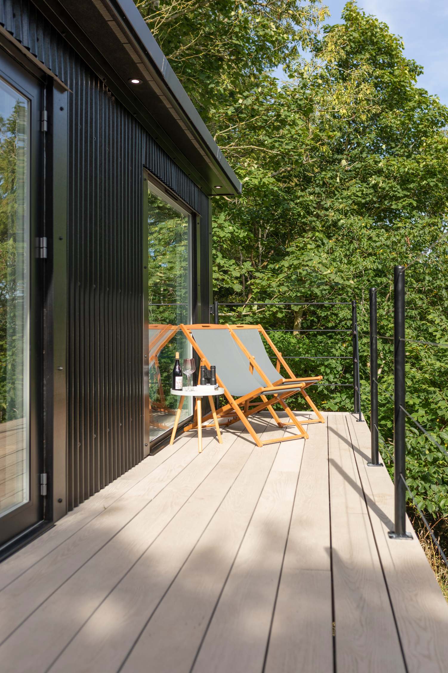 Two orange-framed deckchairs face a round table set with a wine bottle, glasses and a book, on a deck beside glazed doors, trees behind.