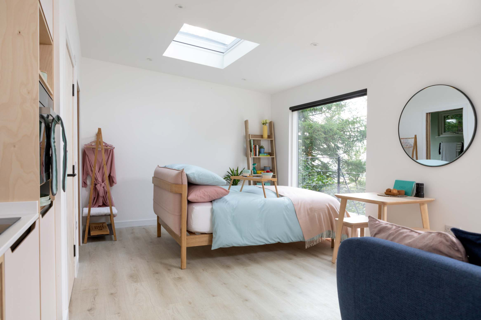 A bedroom with a skylight above the bed, a round mirror on the wall and a small desk and stool by the window.