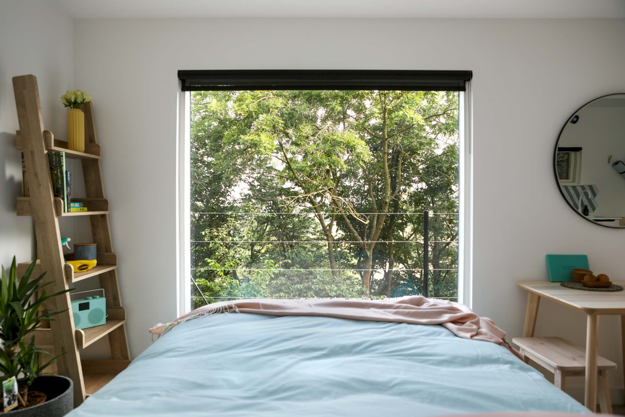 A double bed with a pale blue duvet and a pink throw faces a floor-to-ceiling window full of trees, a ladder shelf of plants and books beside it and a round mirror above a small desk.