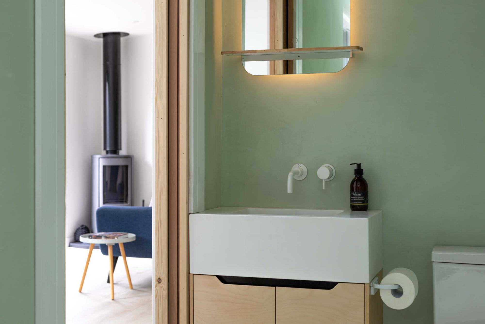 A wall-mounted basin with plain taps sits below a lit mirror, with a wood burning stove and blue armchair visible through the open door beyond.