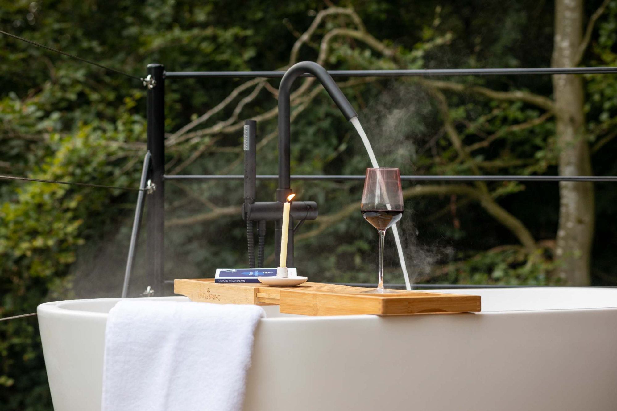 A steaming tap fills a freestanding bath whose rim holds a wooden tray with a lit candle and a glass of red wine, trees behind.
