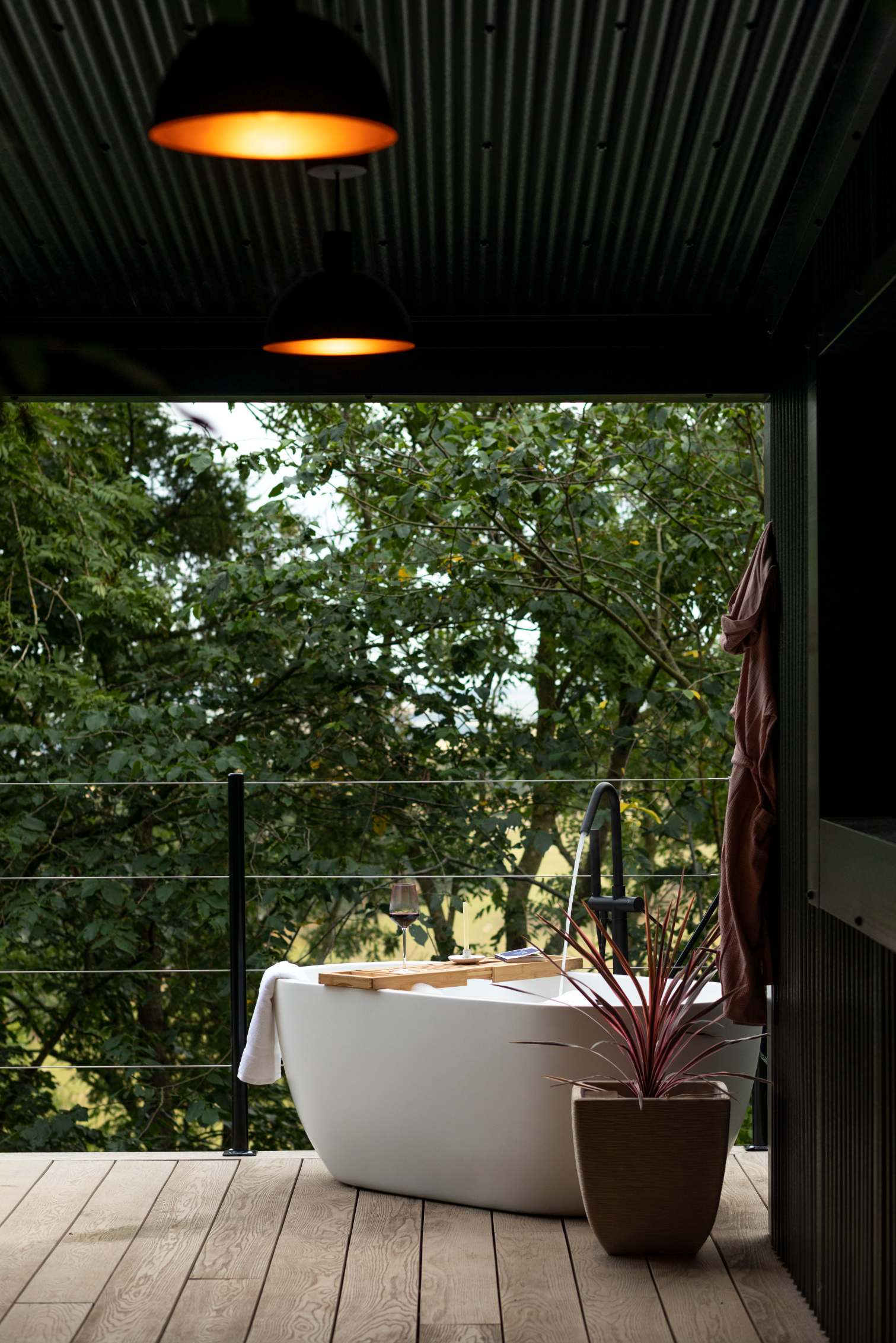 An outdoor bath stands under a dark pitched roof hung with two orange dome lights, open to a view of trees and hills, a robe on a hook to the side.