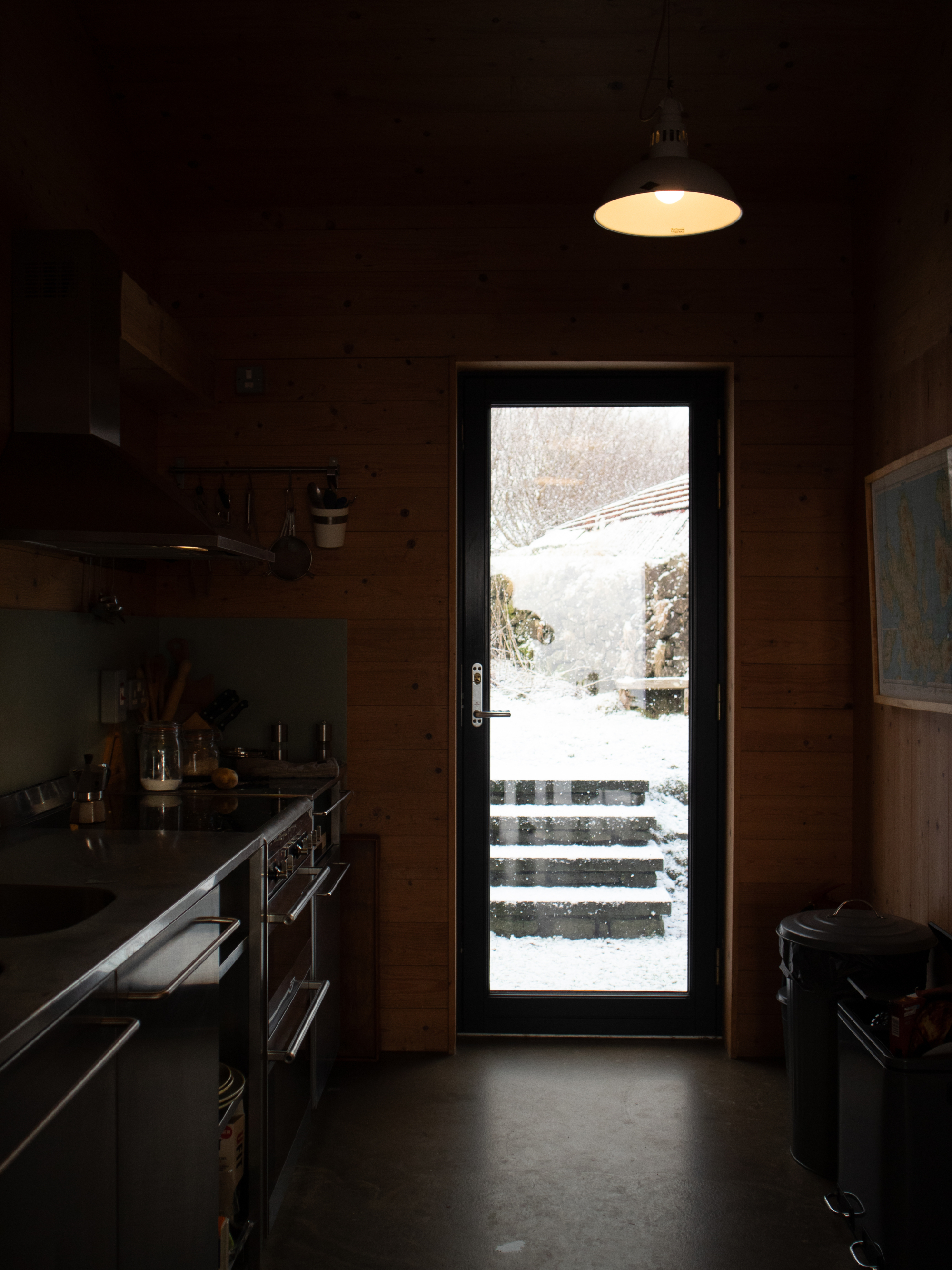 A glazed door lets snow-covered steps and a woodshed into view, a stainless steel kitchen counter in the foreground of the dark room.