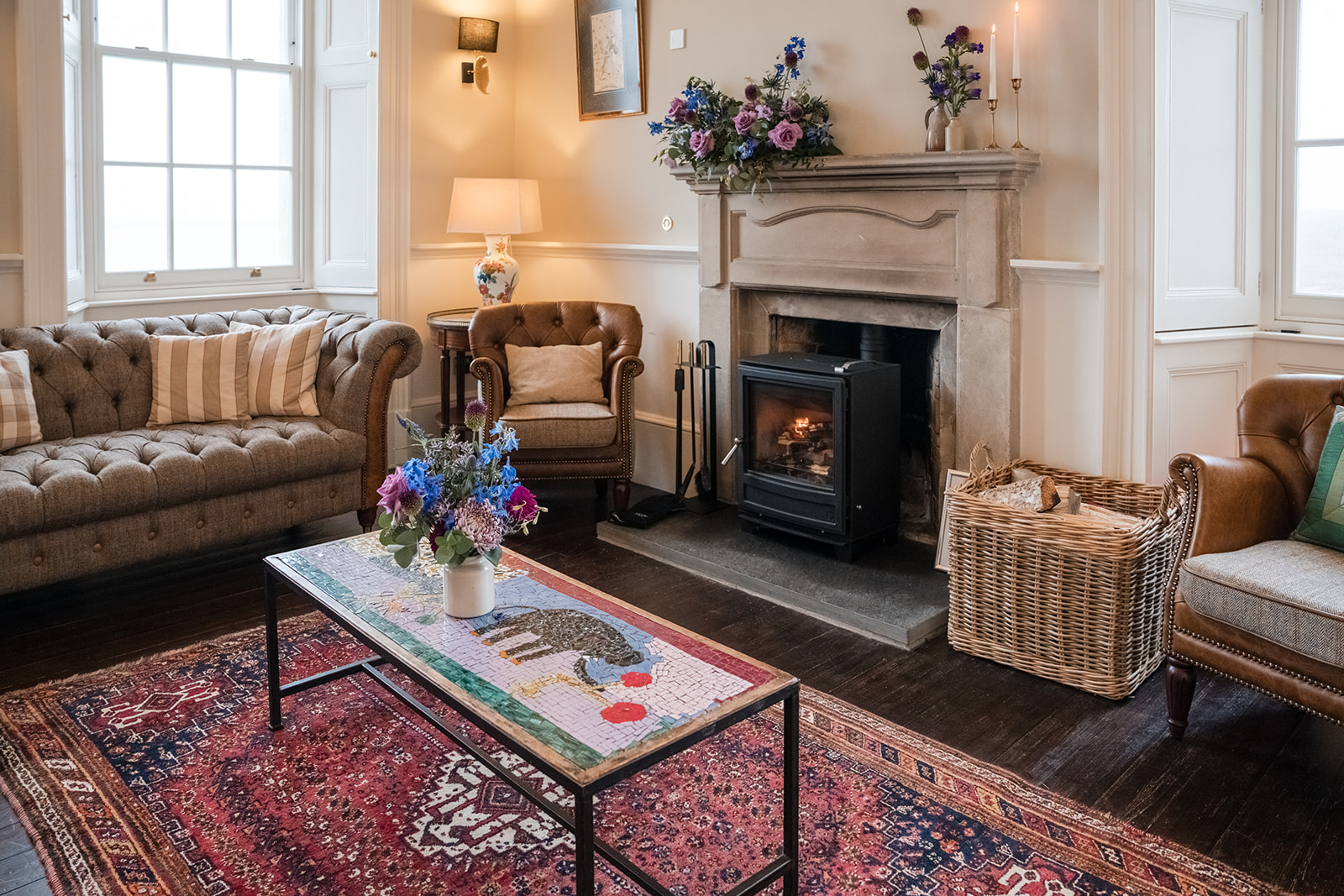 A wood burning stove burns beneath a stone mantel dressed with flowers, a wicker log basket and leather armchair to the side.