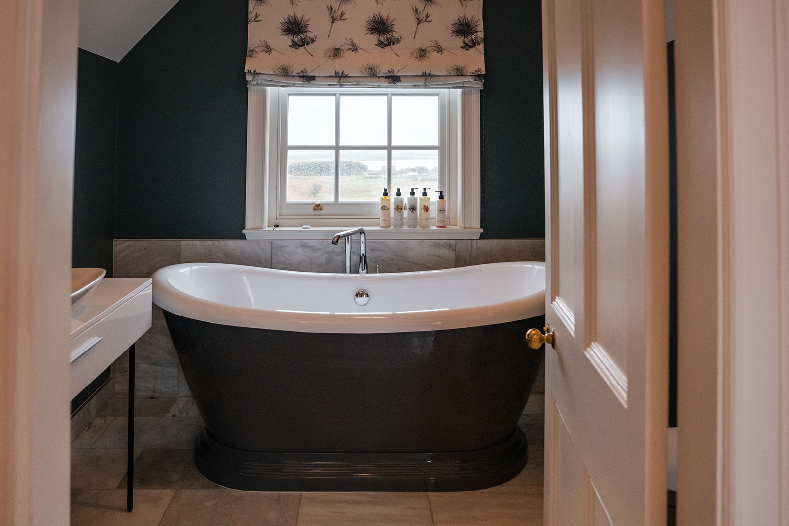 A dark freestanding bath stands before a window with a floral blind, toiletries lined on the sill.