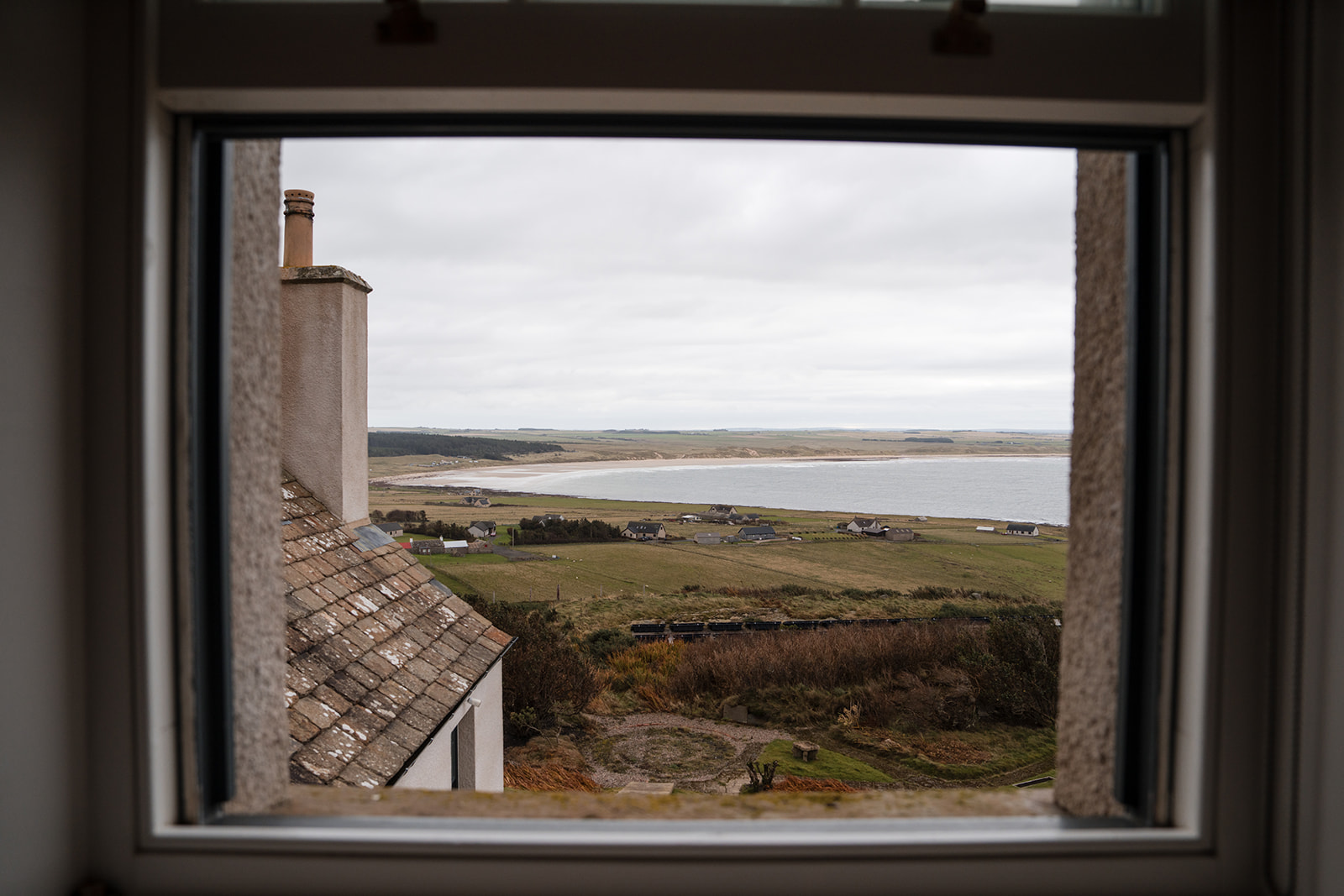 A stone chimney and slated roof are seen through a window, fields and a sea loch stretching to the horizon beyond.