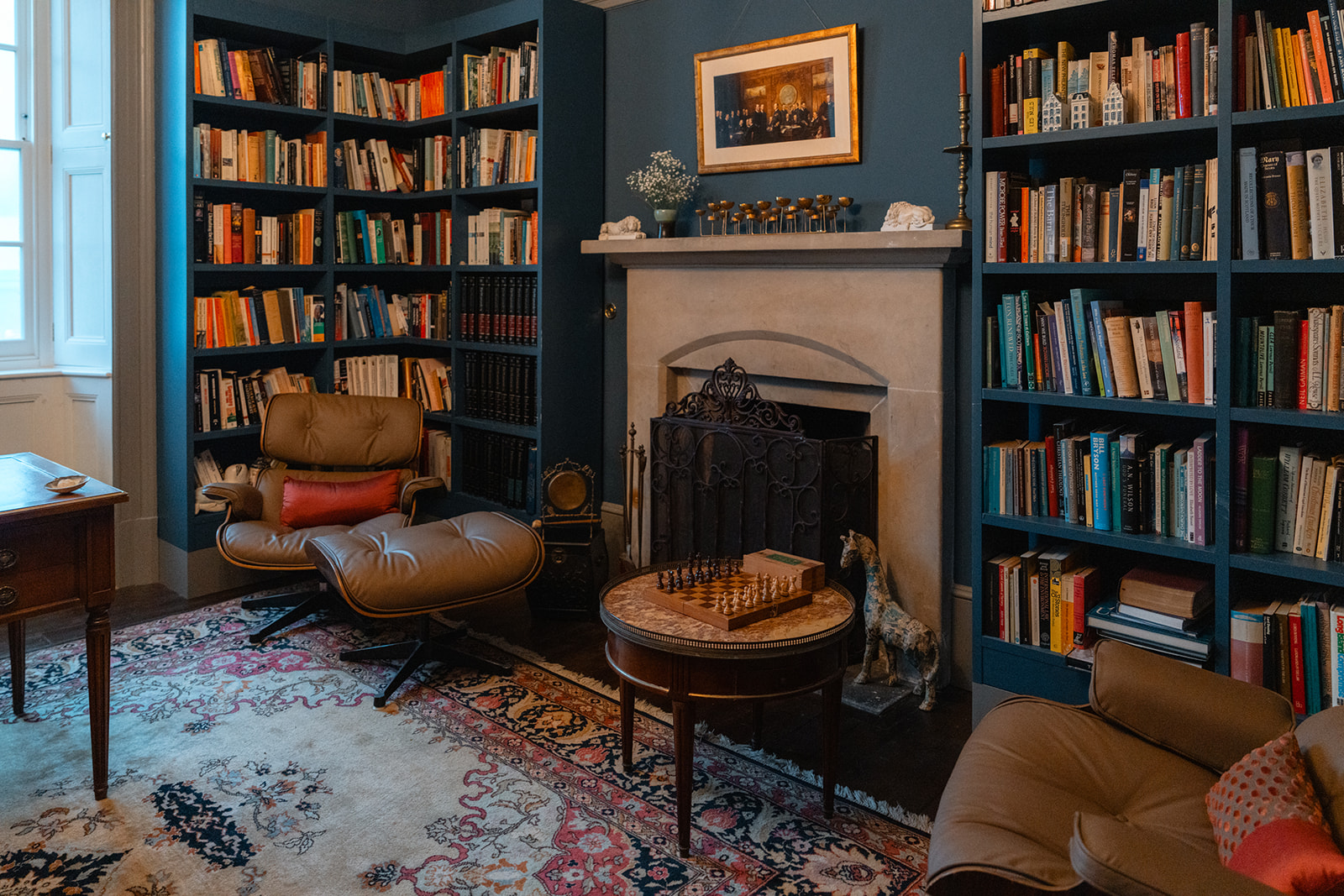 A leather chair and footstool sit beside a small table set with a chess board, bookshelves lining the walls around a fireplace.