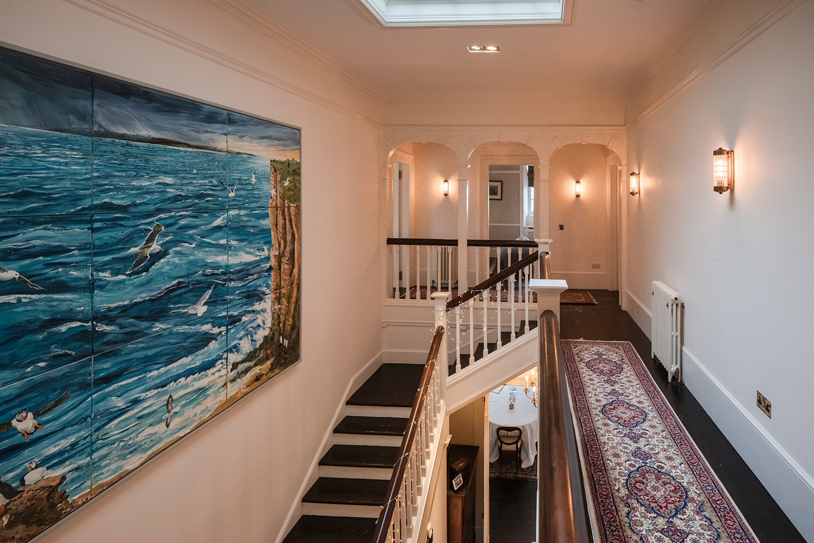 A large painting of a coastal scene hangs on a landing beside an arched doorway, a patterned runner leading down the stairs.
