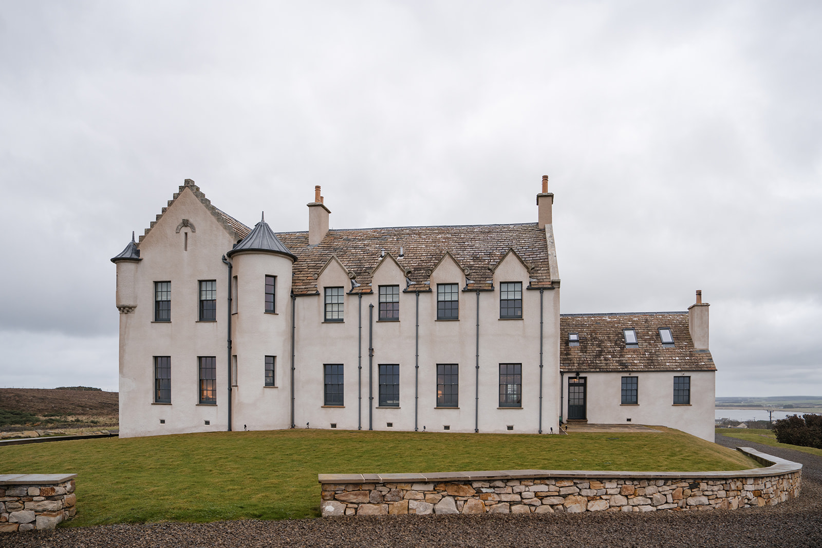 A turreted white mansion with stepped gables and tall chimneys stands behind a low stone wall on a lawn.