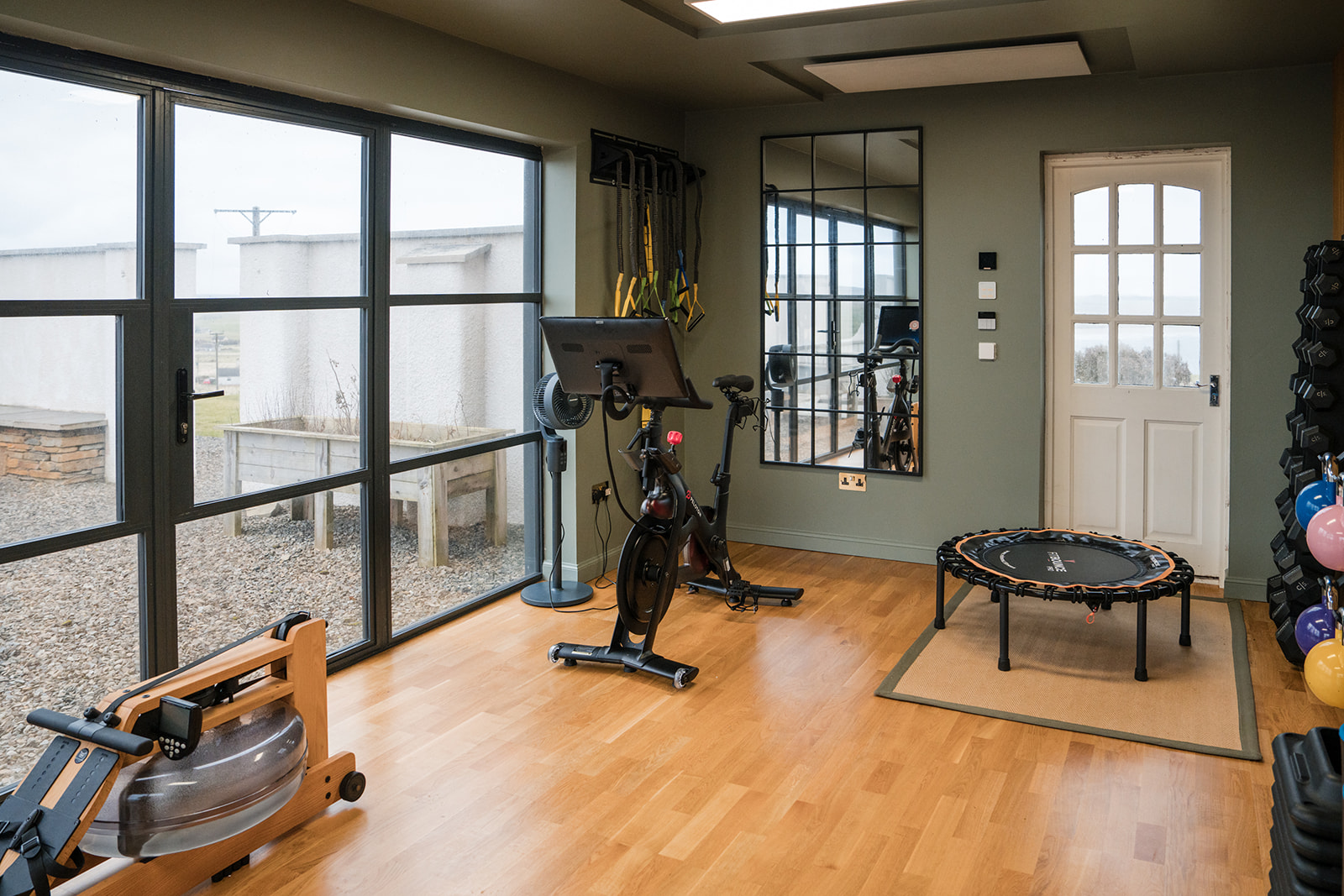 A rowing machine, stationary bike and mini trampoline are arranged in a gym beside full-height windows overlooking a courtyard.